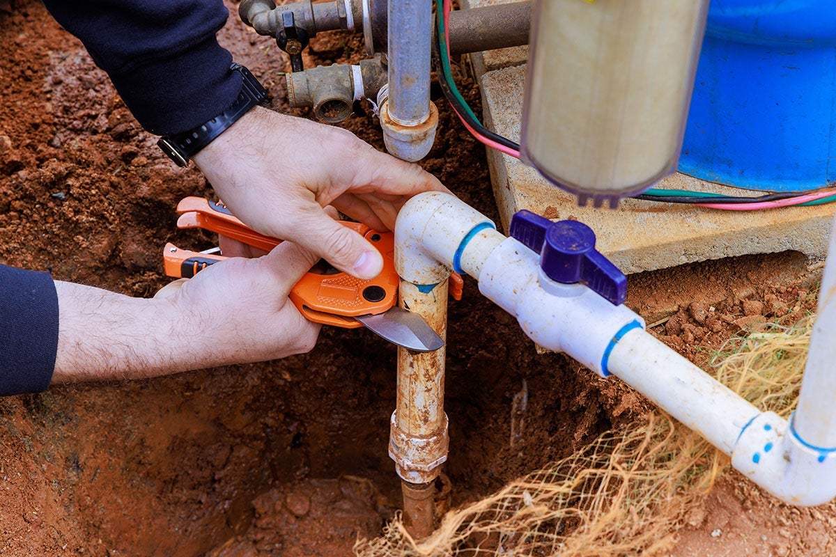 Plumber using pipe cutters to work on an outdoor water line with a ball valve and various pipe connections